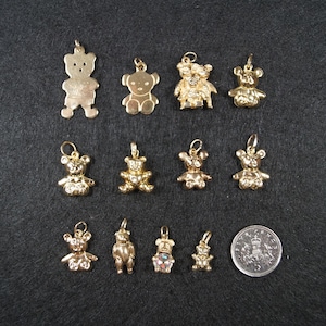 May include: A collection of eleven gold-toned metal teddy bear charms in various sizes and designs, some featuring textured surfaces or embedded stones, arranged alongside a circular silver-colored coin for scale on a dark gray surface.