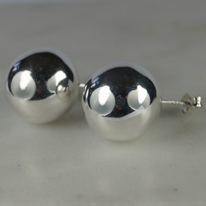 May include: A pair of polished, silver-coloured ball stud earrings. The earrings are round and reflective, with a smooth, shiny surface. The posts and backs are also silver-toned. The earrings are displayed on a white surface.