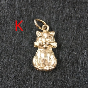 May include: A gold-toned metal charm in the shape of a sitting cat with textured fur details and a small circular jump ring at the top for attachment. The letter K is printed in red to the left of the cat charm.