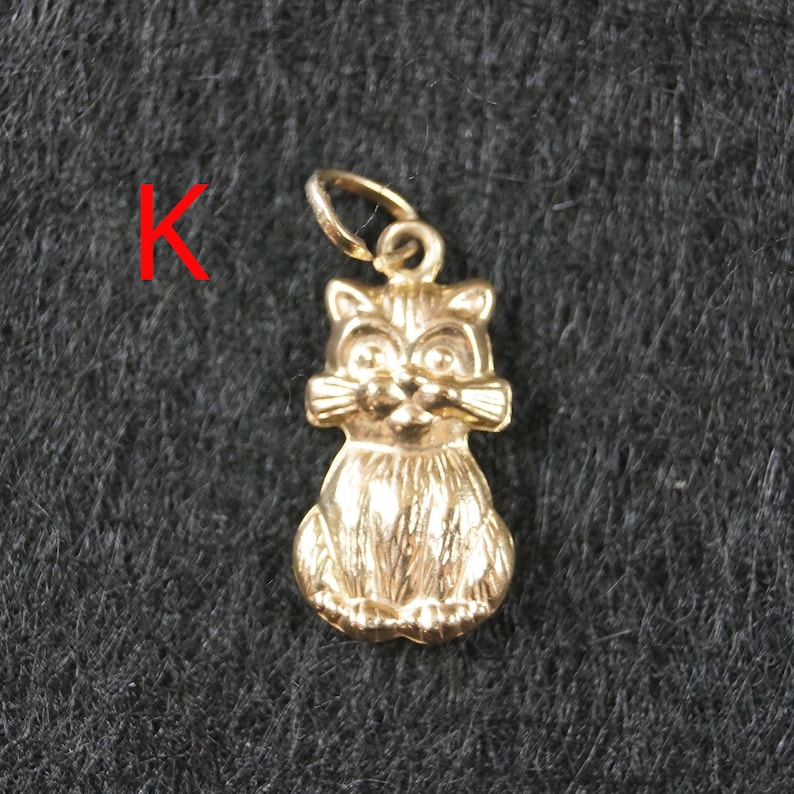 May include: A gold-toned metal charm in the shape of a sitting cat with textured fur details and a small circular jump ring at the top for attachment. The letter K is printed in red to the left of the cat charm.