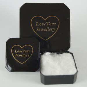 May include: A black jewelry box with a hinged lid and a separate smaller black lid, both featuring a gold heart outline and the text Love Your Jewellery. The open box contains a white cotton insert for holding small items securely inside.