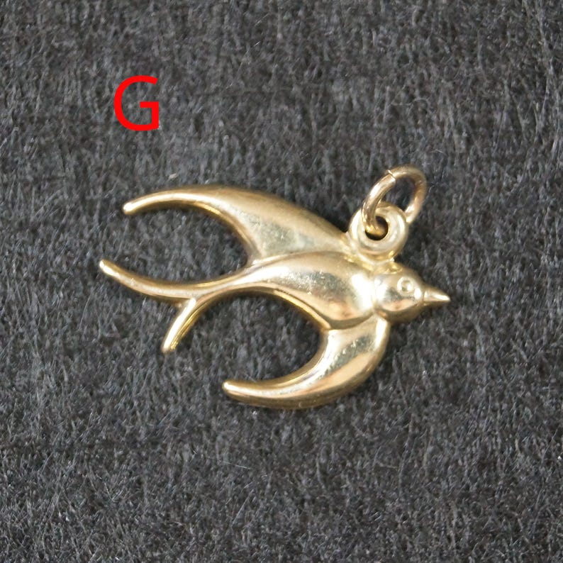 May include: A gold-toned metal charm in the shape of a flying swallow bird with outstretched wings and a small circular jump ring attached to the top. The letter G is printed in red in the upper left corner of the image.