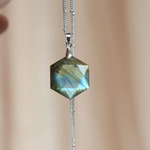 May include: A hexagonal pendant necklace with a silver chain. The pendant is a faceted gemstone with blue, green, and gold hues. The necklace is displayed against a soft, neutral background.
