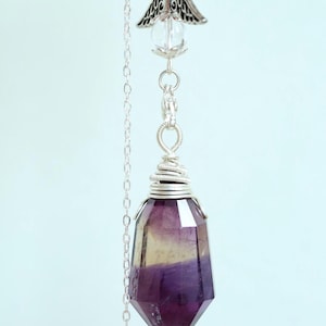 May include: A pendulum featuring a faceted purple and yellow crystal point, suspended by a silver chain. The chain has clear crystal beads and a decorative silver angel wing charm. The crystal is approximately 5 cm long.