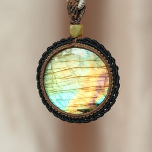 May include: A handcrafted pendant necklace featuring a large, round labradorite stone with iridescent colours of blue, yellow, and green. The stone is set in a macrame design with black and brown cord, and a square green bead. The necklace cord is brown and silver.