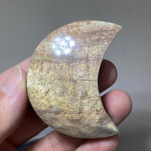May include: A crescent moon-shaped sunstone carving featuring a polished surface with a shimmering, metallic golden-orange aventurescence. The stone displays internal reflective inclusions that create a sparkling effect across its curved form.