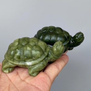 以下が含まれることがあります： 滑らかで磨かれた表面を持つ、石を彫って作られた2体のカメの置物。手前のカメは明るいオリーブグリーンで鉱物の筋が見え、後ろのカメは濃いフォレストグリーンで均一な色合いをしています。