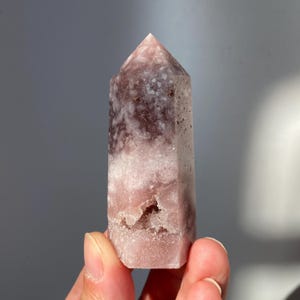 Può includere: Una torre di cristallo rosa e bianca con la parte superiore a punta. Il cristallo ha un aspetto a strati con un mix di sezioni opache e traslucide. La base del cristallo ha una piccola cavità. Il cristallo è tenuto in una mano.