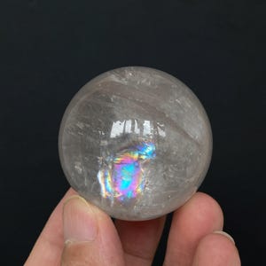 May include: A clear, spherical quartz crystal ball featuring internal fractures and inclusions that create a vibrant, multi-coloured rainbow refraction effect within the centre of the stone, showcasing natural transparency and light reflection.