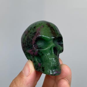 May include: A carved skull figurine made of green zoisite with embedded magenta ruby inclusions. The stone features a polished, smooth surface with natural dark speckling throughout the structure, showcasing deep green and vibrant pinkish-red hues.