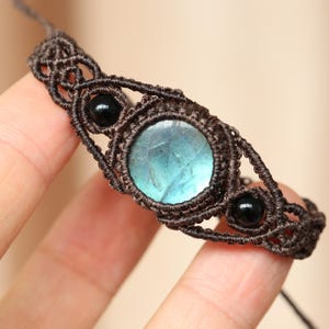 May include: A macrame bracelet with a central, round, iridescent blue-green gemstone, and two small black beads. The bracelet is made with dark brown cord, displaying detailed knotting. A unique piece of jewellery.