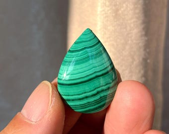 Pode incluir: Uma pedra preciosa de malaquite em forma de gota polida. A pedra apresenta faixas de tons verdes variados, criando um padrão listrado. A pedra é segurada entre dois dedos. Medidas em centímetros.