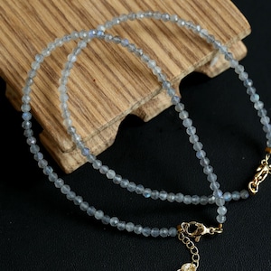 May include: Two delicate necklaces with small, faceted, light blue beads. The necklaces have gold-colored clasps and are displayed on a wooden surface against a dark background. The beads have a subtle shimmer.
