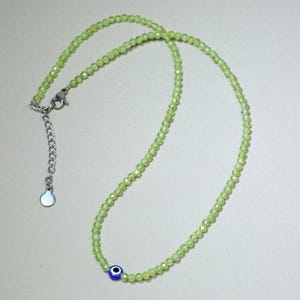 Könnte beinhalten: Eine zarte Halskette mit kleinen, hellgrünen Perlen. Sie hat einen silberfarbenen Verschluss und eine verstellbare Kette. Ein blau-weißer Evil-Eye-Anhänger ist in das Design integriert und setzt einen einzigartigen Akzent.