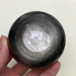 May include: A polished, spherical silver obsidian stone featuring a smooth, reflective surface with a distinct metallic sheen. The dark, circular mineral displays a gradient of deep black tones transitioning into a bright, shimmering silver centre.