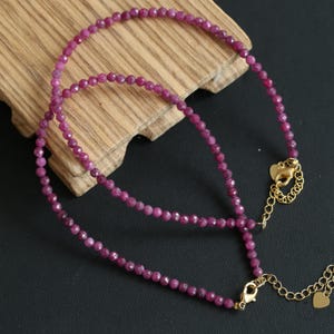 Può includere: Due collane con piccole pietre preziose sfaccettate di colore rosa intenso. Le collane hanno chiusure ed estensioni color oro. Le collane sono esposte su una superficie di legno su sfondo scuro.