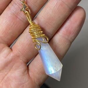 Può includere: Un ciondolo dorato avvolto in filo metallico con un cristallo sfaccettato e iridescente. Il cristallo è di colore bianco e azzurro pallido. Il ciondolo ha una chiusura dorata ed è progettato per essere indossato come collana.