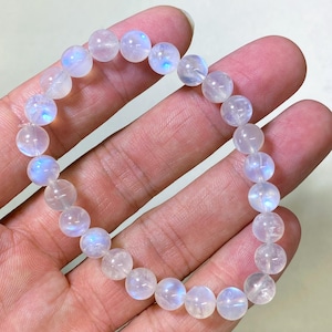 Op de afbeelding: Een armband met ronde, doorschijnende maansteen kralen. De kralen hebben een subtiele iriserende glans, met blauwe en witte flitsen. De armband is geregen op een elastisch koord. De kralen hebben een diameter van ongeveer 0,8 cm.