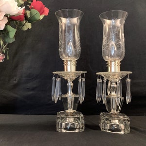 May include: Two clear glass candle holders with etched designs and crystal prisms hanging from the base. The candle holders are on a black background.