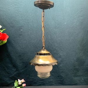 May include: Vintage brass pendant light with a scalloped shade and frosted glass globe. The light fixture hangs from a chain and a decorative ceiling mount. The fixture has a classic, antique design.