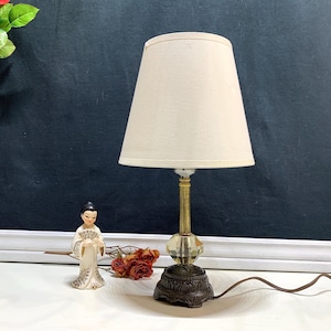 May include: A vintage table lamp with a cream-colored lampshade. The lamp features a brass stem, a faceted crystal accent, and a decorative bronze base. A small figurine and dried flowers are next to the lamp.