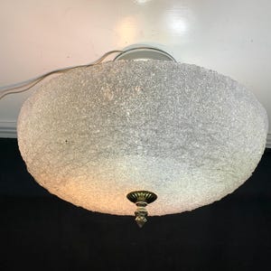 May include: An oval ceiling light with a textured, off-white shade. The fixture features a brass-coloured decorative element and a silver mounting plate. The light fitting is wired for electricity.