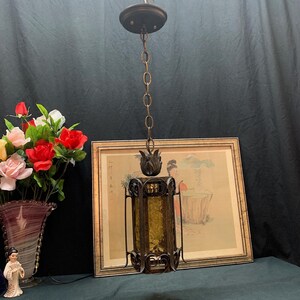 May include: A hanging pendant light fixture featuring a cylindrical amber-colored textured glass shade encased in a dark brown metal frame with scrollwork details, suspended by a matching metal chain from a circular ceiling canopy mount.