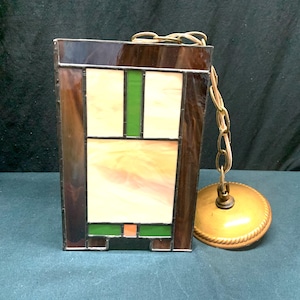 May include: Rectangular stained glass pendant light fixture with a geometric design. The fixture features brown, green, orange, and cream-colored glass panels. A brass-colored chain and ceiling mount are attached.