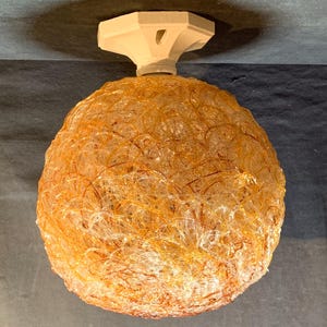 May include: An amber-coloured spherical light fitting with a woven texture. It is suspended from a white, geometric ceiling mount. The design of the light suggests a modern or vintage style. The light's diameter is approximately 30 cm.