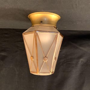 May include: An Art Deco style ceiling light fixture. The light has a gold-colored metal base and trim, with a frosted glass shade. The shade is geometric, with a series of triangles and circles. The light is designed to be mounted flush against the ceiling.