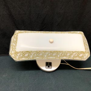 May include: A rectangular wall-mounted light fitting featuring a white frosted glass shade with a gold-coloured decorative border pattern. The fitting includes a central white knob and a white base with a built-in black electrical socket.