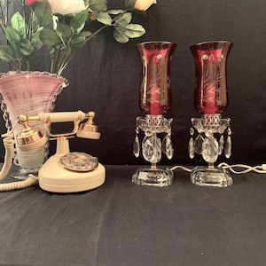 May include: Two vintage crystal candle holders with burgundy glass candle shades and crystal teardrop accents. A white rotary phone with gold accents is also in the image.