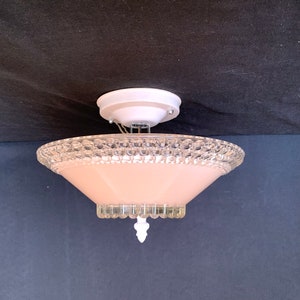 May include: Vintage ceiling light fixture with a white canopy and a pink glass shade. The shade has a clear, textured rim and a decorative white finial. The light fixture is designed to be mounted on the ceiling.