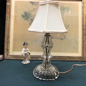 May include: A clear glass table lamp with a white lampshade. The lamp has a decorative, faceted base and stem. A small porcelain figurine of a woman in a traditional outfit stands nearby. The background features a framed Asian-style print.