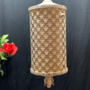 May include: Cream-colored vintage hanging lamp with a cylindrical shape. The lamp features an intricate, openwork design with a floral pattern. The top and bottom edges are adorned with decorative trim. A red rose is visible in the background.