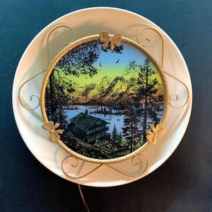 May include: Vintage decorative plate with a landscape scene featuring a lake, mountains, and trees. The image is framed with a gold-colored metal frame and bow accents, set within a white, round plate.
