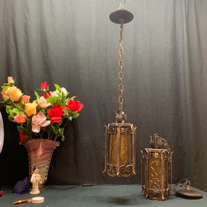 May include: Two cylindrical pendant lights featuring dark bronze-toned metal frames with intricate scrollwork and textured amber glass panels. Each light is suspended by a matching metal chain and includes a decorative metal cap at the top.