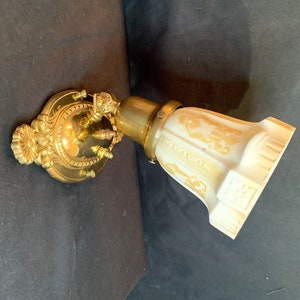 Peut inclure: Applique murale en laiton doré orné avec un abat-jour en verre crème. L'abat-jour présente un motif floral doré. L'applique a une plaque arrière décorative et un bras en laiton. Le luminaire est conçu pour une seule ampoule.