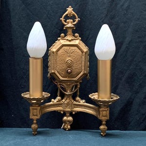 May include: A gold-toned metal wall sconce featuring an ornate central octagonal plate with textured detailing and a decorative finial. The fixture holds two vertical candle-style light sockets topped with white, flame-shaped frosted glass bulbs.