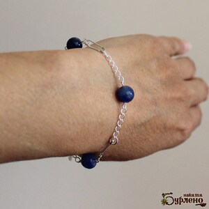 May include: A silver chain bracelet with three dark blue, spherical beads. The bracelet is fastened with a safety pin and is worn on a wrist. The background is a neutral color.