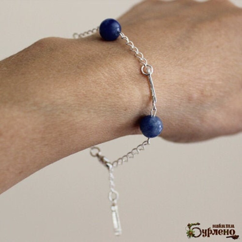 May include: A silver chain bracelet featuring two dark blue, spherical beads. The bracelet has a delicate chain design and a clasp closure. The beads are a deep, rich blue color, adding a pop of color to the silver chain.