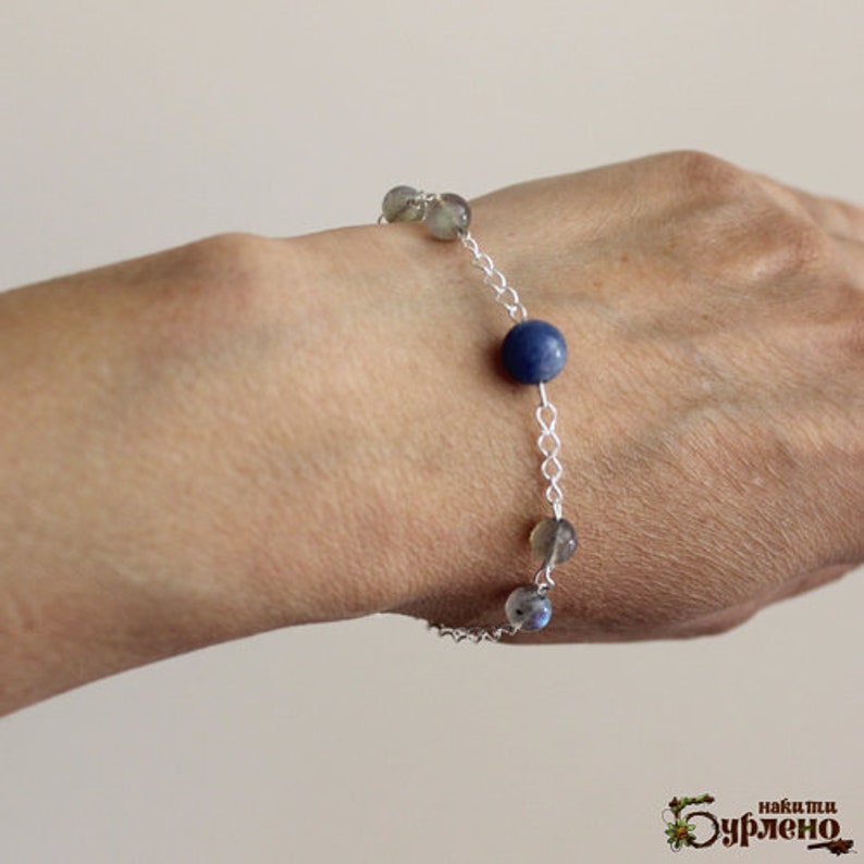 K&ouml;nnte beinhalten: Ein silbernes Kettenarmband mit blauen und grauen Edelsteinperlen. Das Armband befindet sich am Handgelenk einer Person.