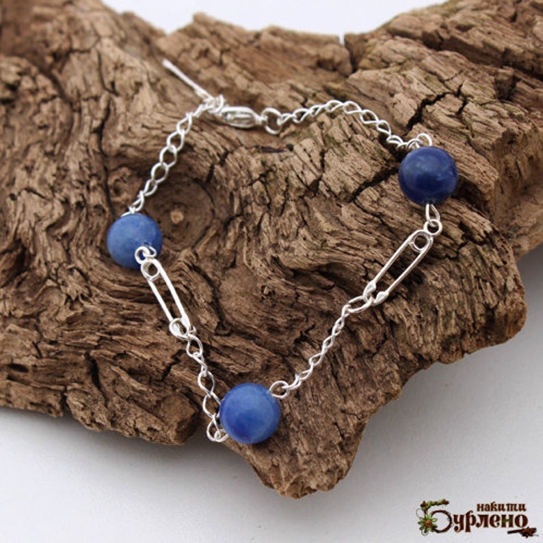 May include: A silver chain bracelet featuring three blue spherical beads and silver safety pin accents. The bracelet is displayed on a piece of textured wood.