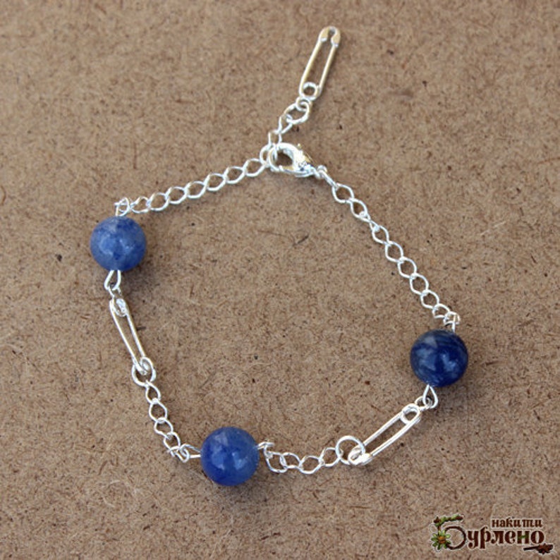 May include: A silver bracelet featuring three blue spherical beads. The bracelet is made with a chain and safety pin links. The clasp is silver. The bracelet is on a brown surface.