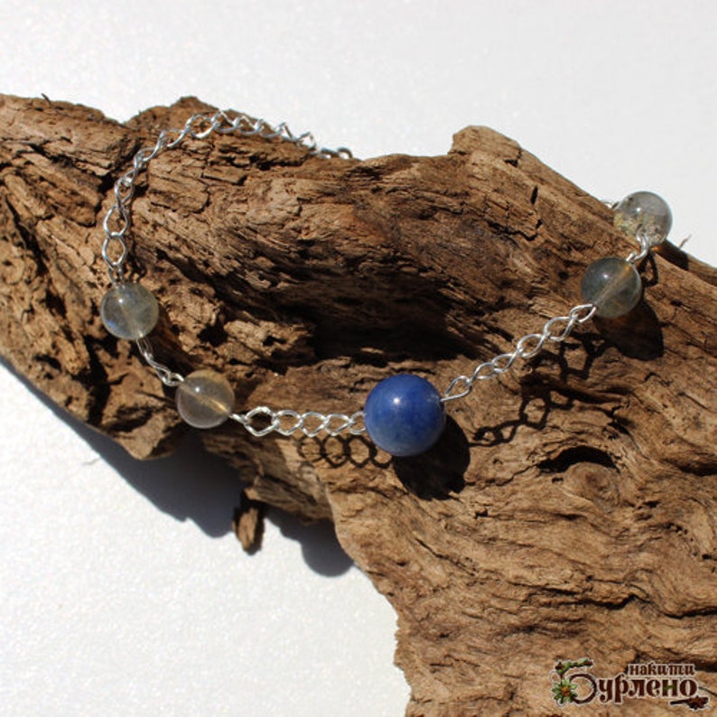 K&ouml;nnte beinhalten: Ein silbernes Kettenarmband mit vier runden Perlen. Die Perlen sind dunkelblau, hellgrau und zwei mittelgrau. Das Armband liegt auf einem St&uuml;ck verwittertem Holz.