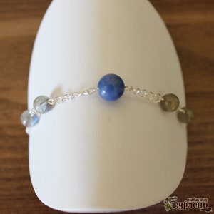 K&ouml;nnte beinhalten: Ein silbernes Kettenarmband mit blauen und grauen Edelsteinperlen. Das Armband hat einen blauen Edelstein in der Mitte und graue Edelsteine ​​auf beiden Seiten.