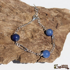 May include: A silver chain bracelet with three blue gemstone beads. The bracelet is set against a textured, brown wooden surface. The bracelet's design incorporates chain links and decorative metal elements.