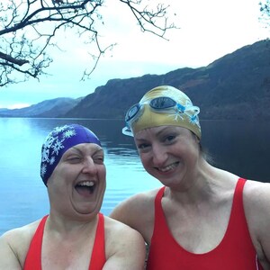 May include: Two women wearing red swimsuits and swim caps with snowflake designs are smiling at the camera. They are standing in front of a lake with mountains in the background.
