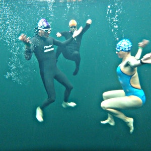 May include: Three people wearing swim caps and swimsuits are underwater. One person is wearing a black wetsuit and a purple and white swim cap. Another person is wearing a blue swimsuit and a blue and white swim cap. The third person is wearing a black wetsuit and a yellow swim cap.