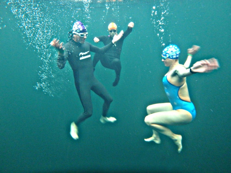 May include: Three people wearing swim caps and swimsuits are underwater. One person is wearing a black wetsuit and a purple and white swim cap. Another person is wearing a blue swimsuit and a blue and white swim cap. The third person is wearing a black wetsuit and a yellow swim cap.